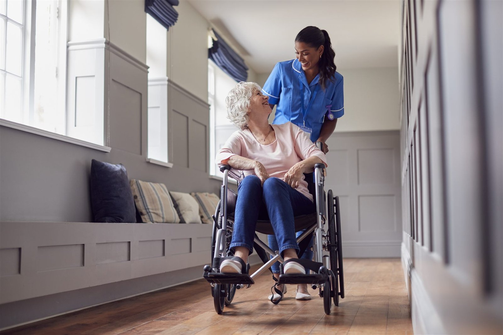 A smiling nurse chatting warmly with an older lady as she pushes her wheelchair along a bright hallway